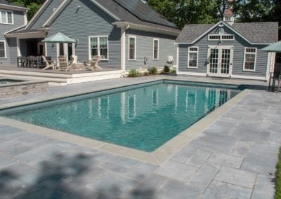 Backyard with a rectangular swimming pool designed by a top-rated pool company and a stone patio, adjacent to a gray house with white trim.