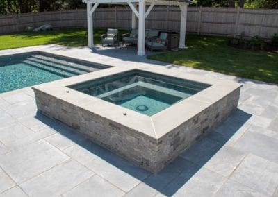 An in-ground spa adjacent to a swimming pool, expertly crafted by a top pool company, with a backyard patio and pergola in the background.
