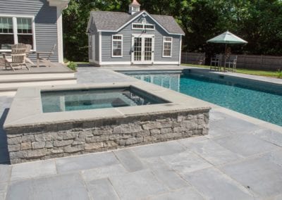 A residential backyard featuring an in-ground swimming pool with an attached hot tub, designed by a premier pool company, surrounded by a stone patio and a pool house in the background.