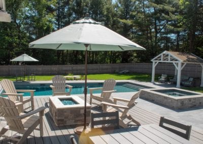 A poolside patio designed by a renowned pool company, featuring Adirondack chairs, umbrellas, and a hot tub on a sunny day.