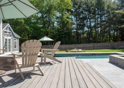 A sunny poolside patio designed by a renowned pool company, featuring adirondack chairs and a view of a landscaped backyard.