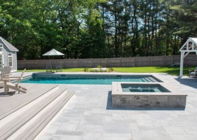 Backyard with a swimming pool designed by a pool company, fire pit, and seating area amidst greenery on a sunny day.