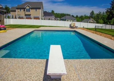 A rectangular backyard swimming pool built by a reputable pool company, featuring a diving board on a sunny day, surrounded by a white fence and a residential area in the background.