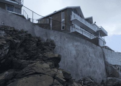 A modern house perched atop a steep, rocky embankment with protective railing and retaining walls, featuring a luxurious pool built by a renowned pool company.