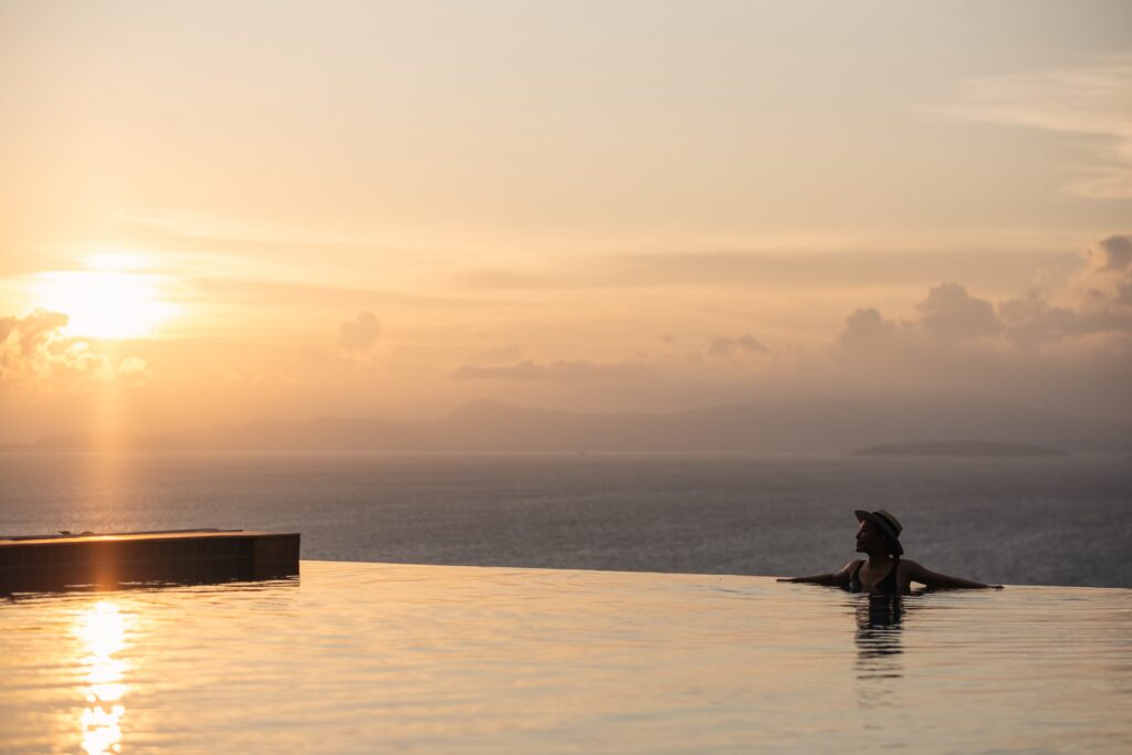 Person in a hat relaxes in a negative edge pool, gazing at a serene ocean view during sunset, with soft clouds and distant mountains in the background.