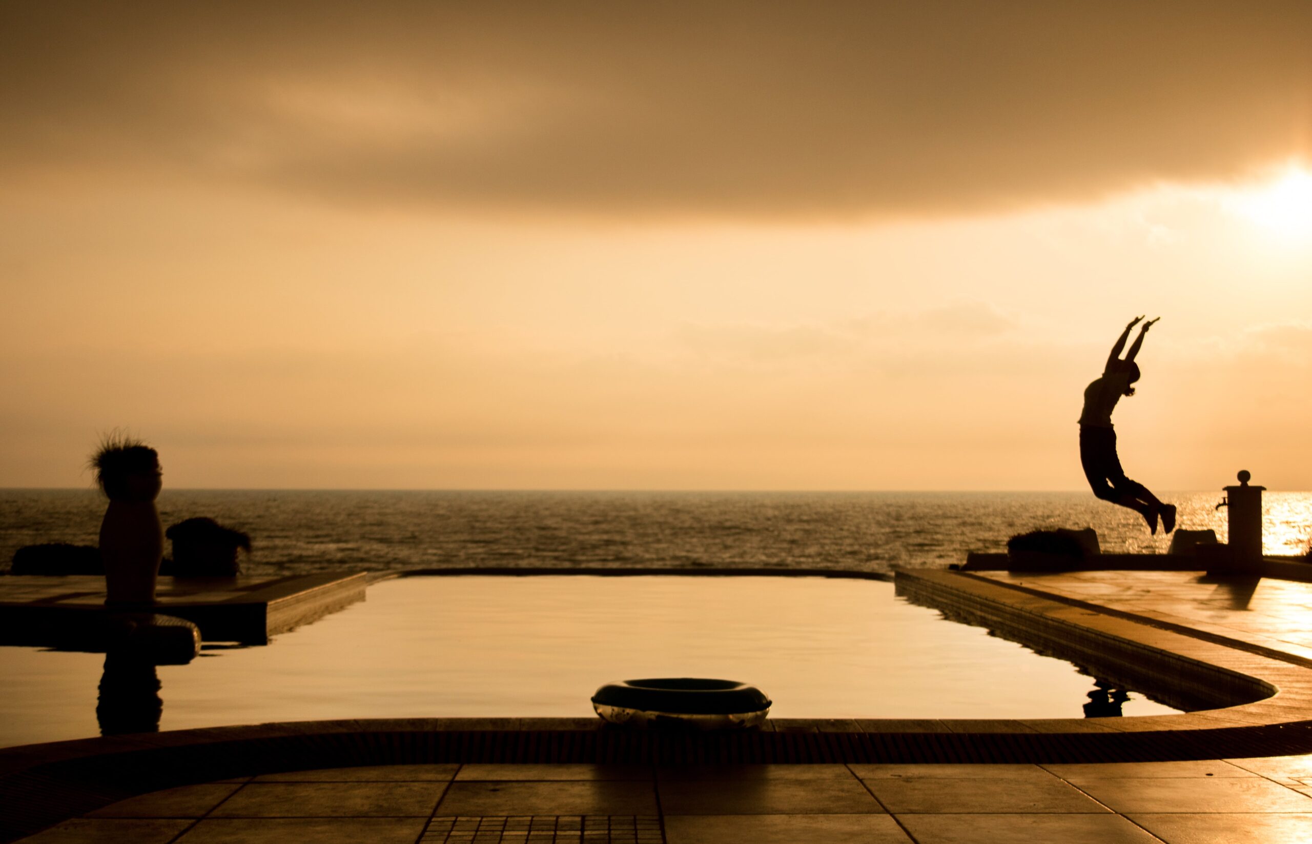 A person is seen in mid-air, jumping into a negative edge pool at sunset with the ocean in the background. Another person stands on the poolside, watching.