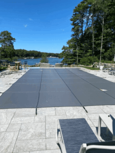 A swimming pool with a gray safety cover, signaling pool closing season, is surrounded by a stone patio with lounge chairs and offers a scenic view of the lake and trees in the background.