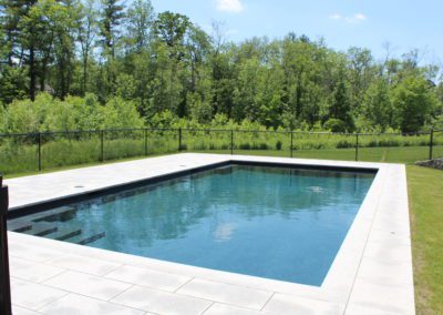 Rectangular outdoor swimming pool with clear blue water, created by a skilled pool builder, bordered by a patio and surrounded by a lush green lawn and trees under a clear sky.