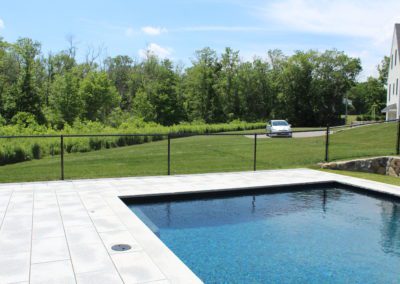 A backyard swimming pool built by a reputable pool company, with a paved deck and a black fence overlooking a wooded area on a sunny day.