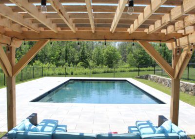 Wooden pergola overlooking a rectangular swimming pool constructed by a top-rated pool company, with loungers on a patio and a manicured lawn in the background.