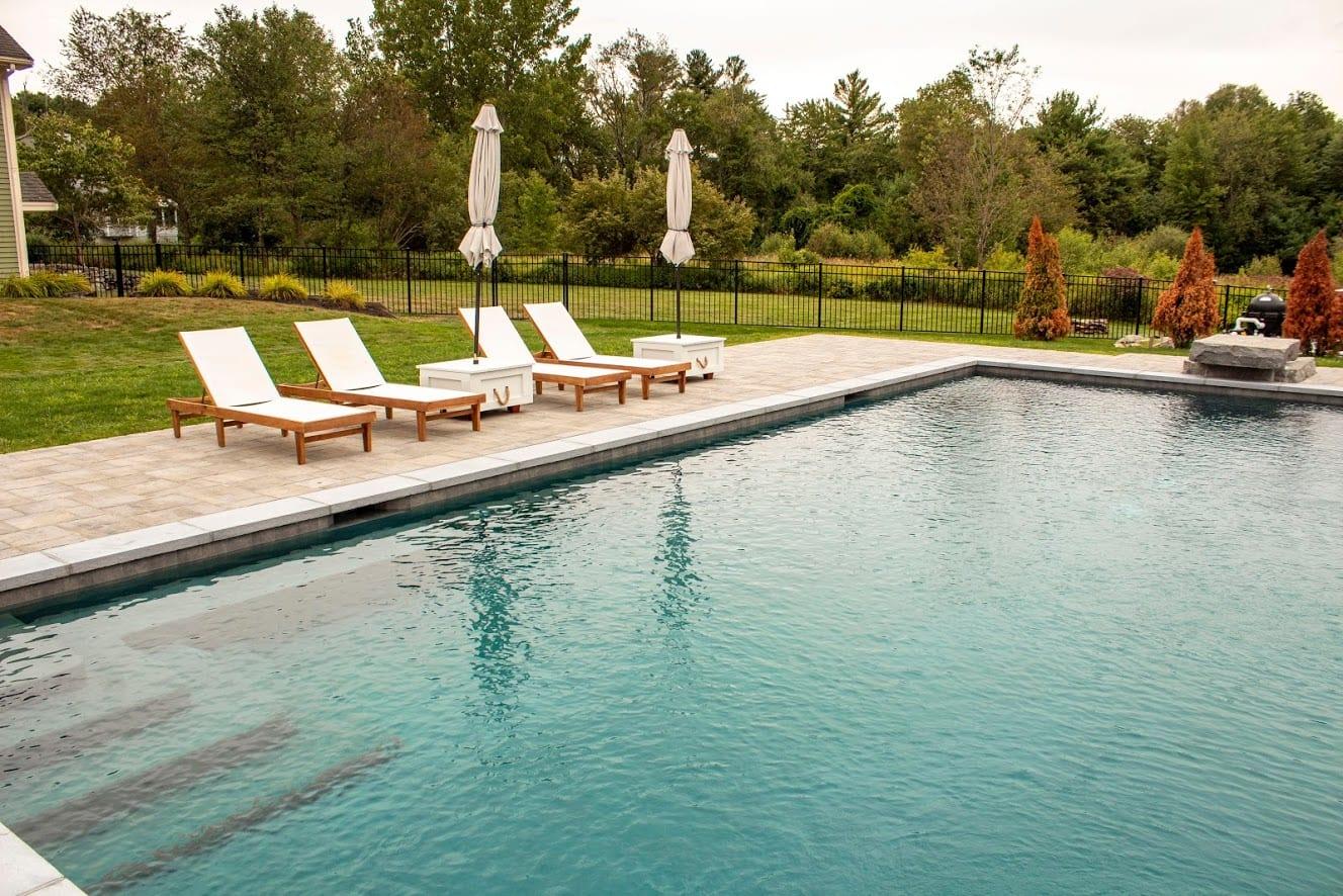 A rectangular gunite pool with four white lounge chairs and two closed umbrellas on a stone deck, surrounded by grass and trees.