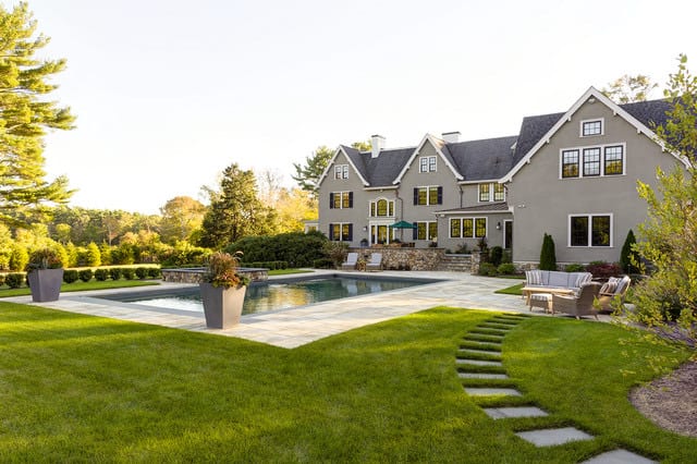 A large house with multiple windows overlooks a rectangular gunite pool, stone patio, and manicured lawn with outdoor seating and potted plants.