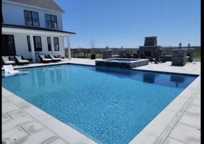 A backyard swimming pool constructed by a professional pool builder with clear blue water adjacent to a white modern house, featuring poolside loungers and a stone fire pit area.