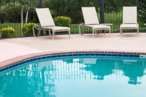 Three empty lounge chairs sit beside an outdoor swimming pool, which looks recently replastered, with a metal fence and green shrubs in the background.
