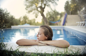 A person with wet hair rests their arms on the freshly replastered edge of an outdoor pool, looking to the side. Trees and a hammock are visible in the background.