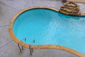 A small, kidney-shaped swimming pool with clear blue water, stone edging, metal ladder, and a rock waterfall feature sits on a concrete patio—recently enhanced with a professional replaster for a fresh and inviting look.