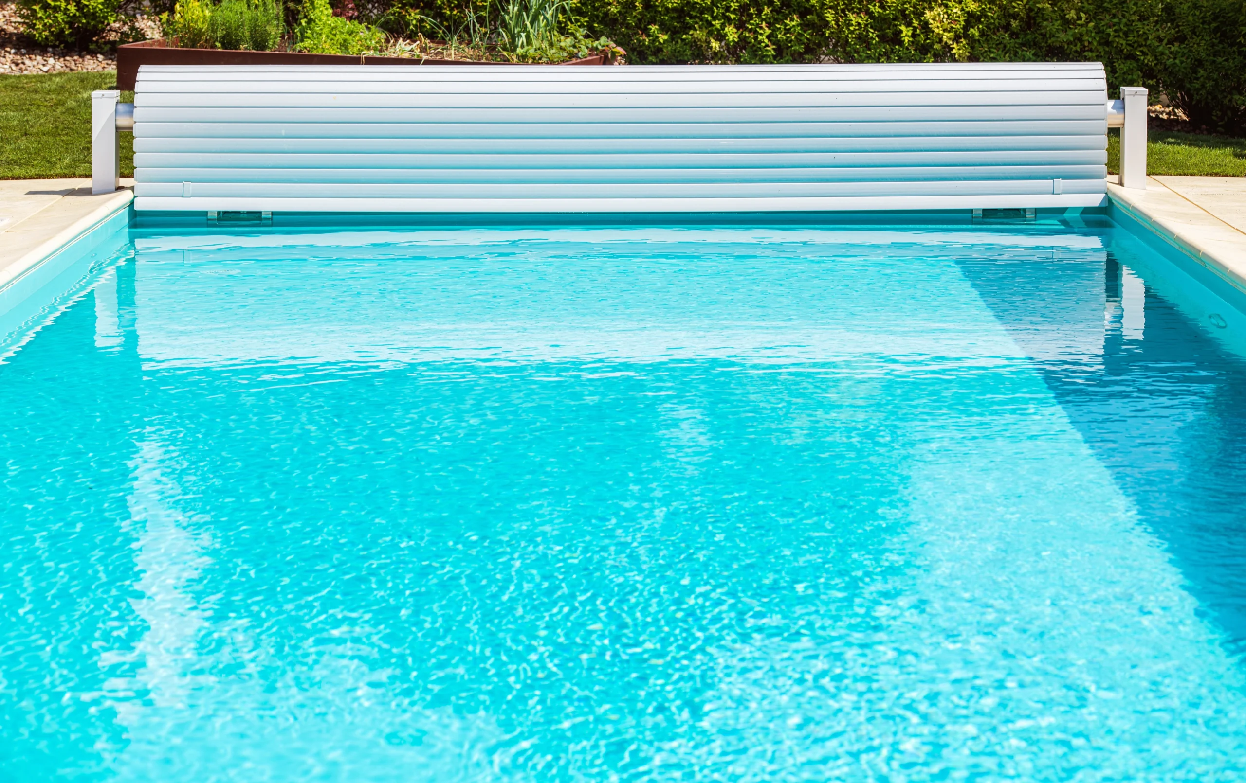 A rectangular outdoor swimming pool with a blue cover partially rolled up at one end, surrounded by a stone deck and greenery in the background, signals the start of opening your pool for the season.