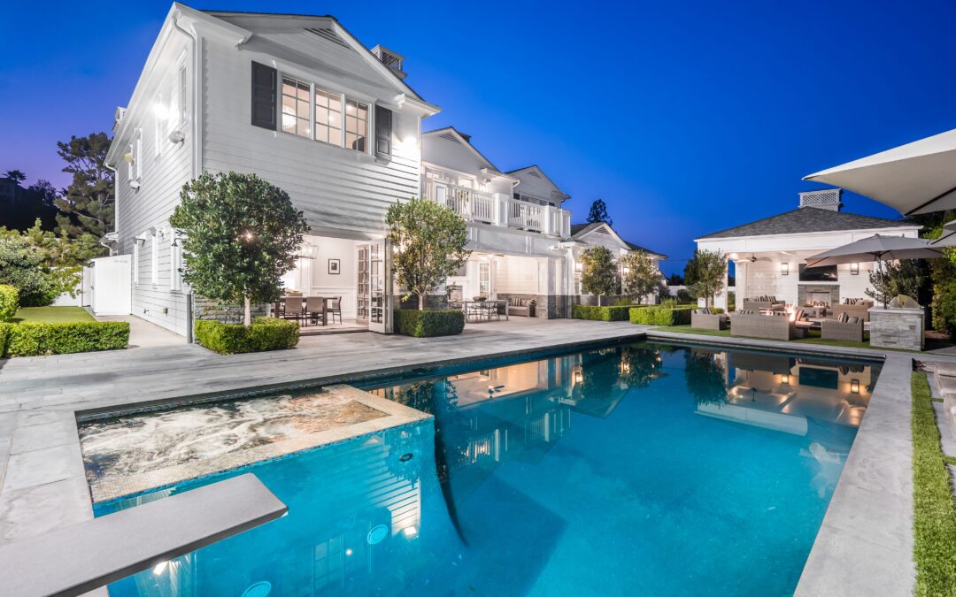 A modern two-story white house with a lit swimming pool in the backyard, surrounded by well-maintained greenery and outdoor furniture, photographed during twilight.