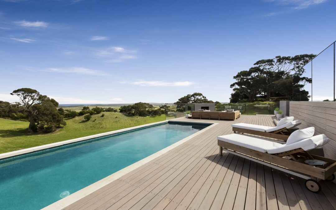 A modern outdoor pool with wooden decking and lounge chairs overlooking a grassy landscape with trees under a bright blue sky.