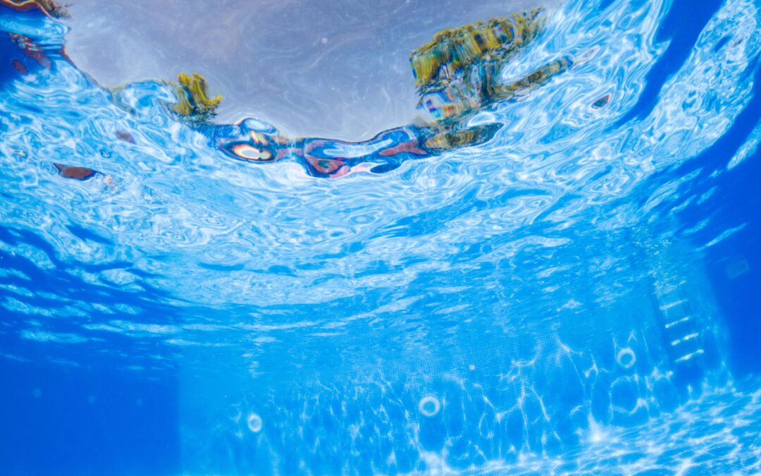 Underwater view of a swimming pool with sunlight reflecting on the rippled surface and blue tiles beneath.