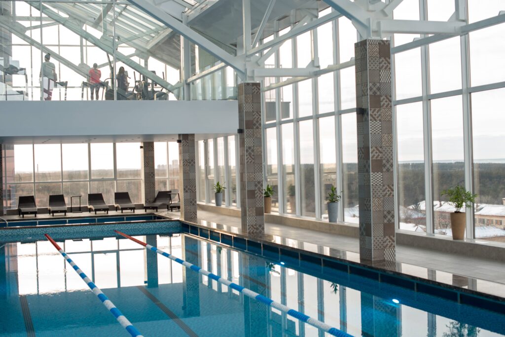 An indoor swimming pool with lane dividers, lounge chairs, and large windows. Several people are visible on an upper level overlooking the pool.