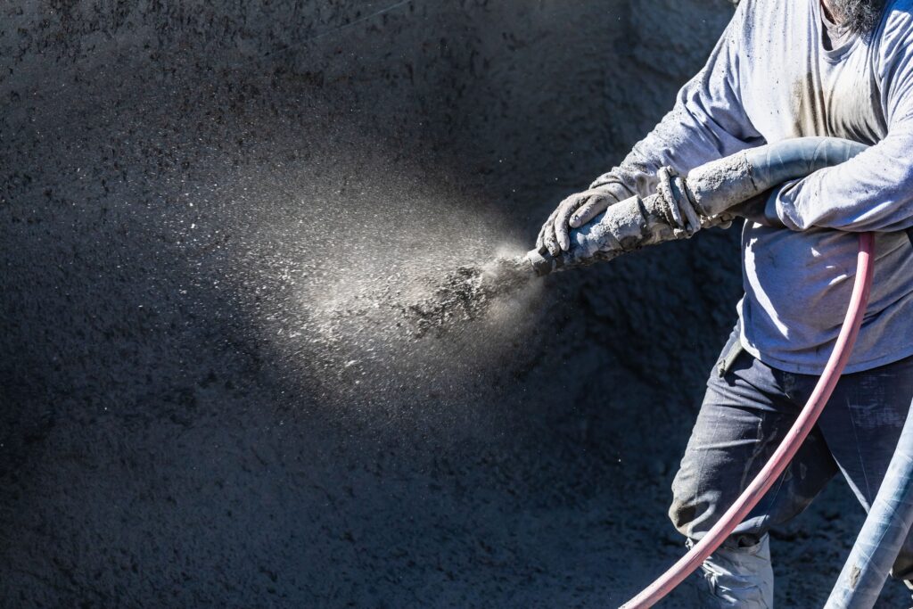 A worker in protective clothing applies sprayed concrete to a surface using a hose, with dust and particles visible in the air.