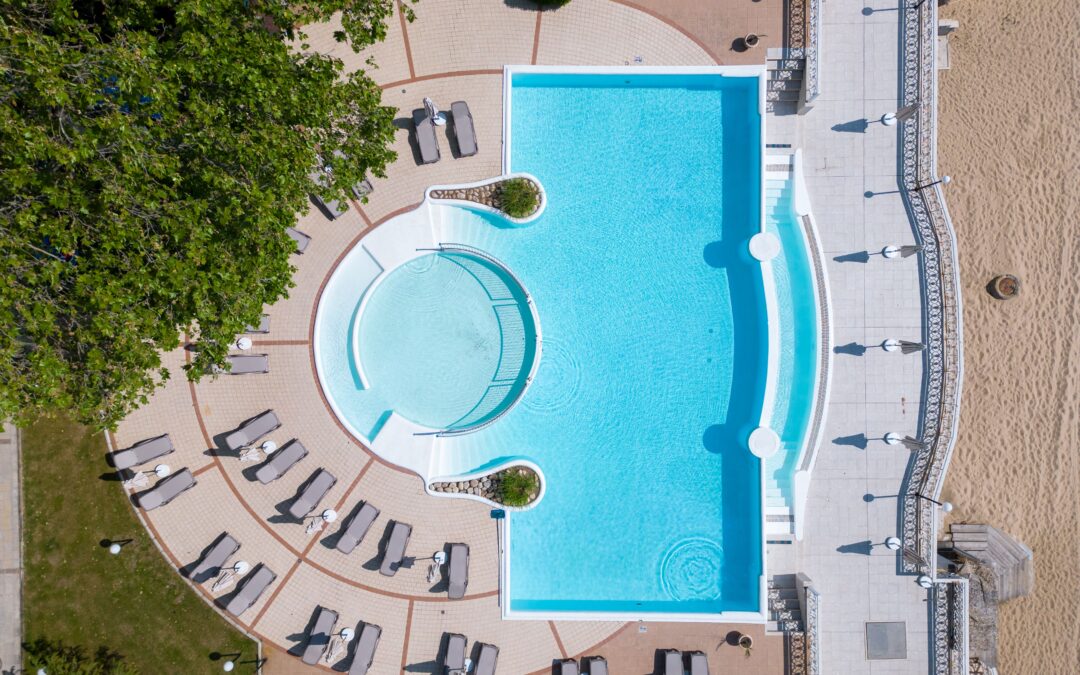 Aerial view of a rectangular swimming pool with lounge chairs arranged around it on a tiled patio, adjacent to greenery and sandy area.