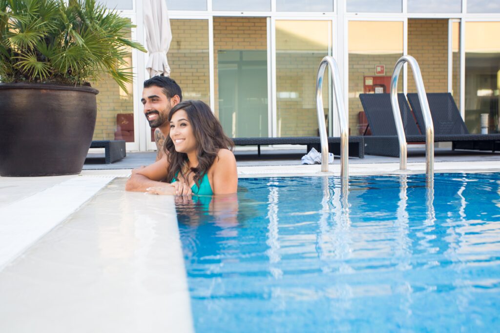 A man and woman in swimsuits are smiling while leaning on the edge of an outdoor swimming pool near lounge chairs and a large potted plant.