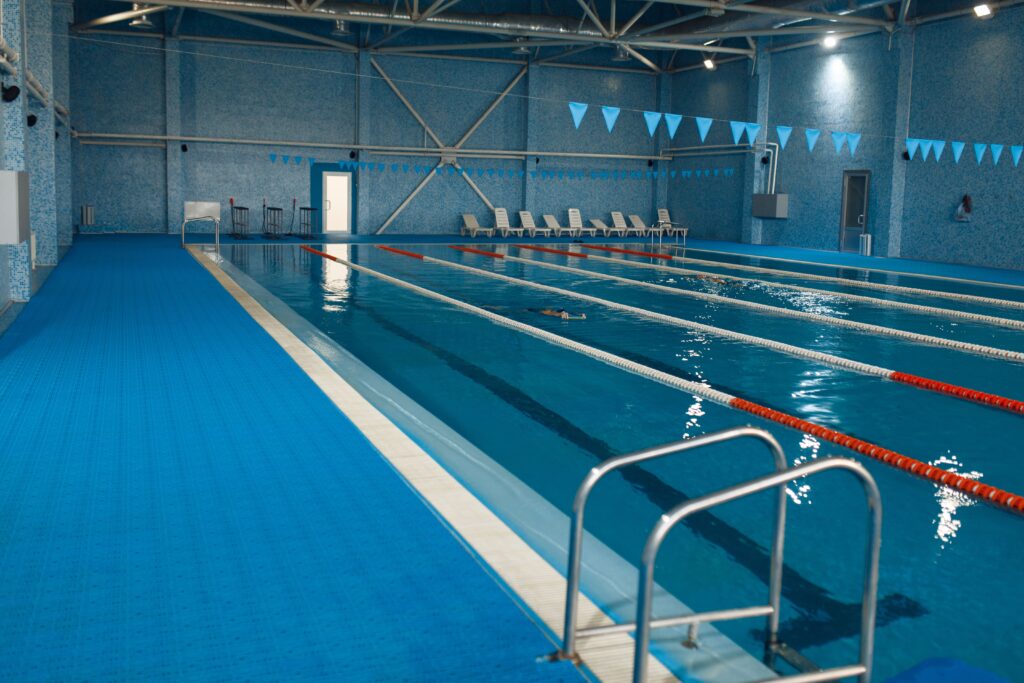Indoor swimming pool with several lanes, blue flooring, lane dividers, pool ladder, and empty lounge chairs along the far wall.