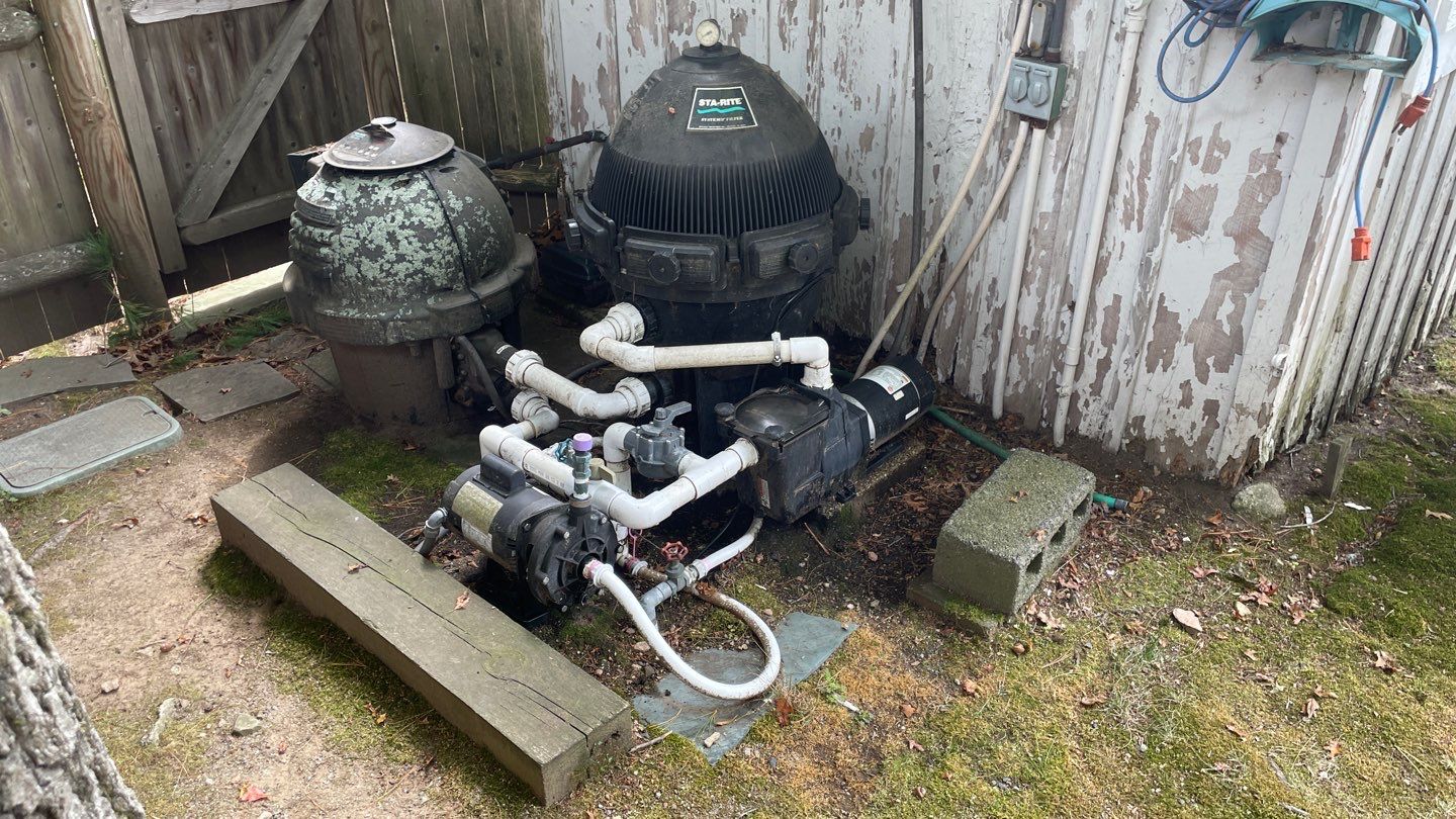 3 Outdoor pool pump and filter system with white PVC piping situated next to a weathered wooden shed and old fence, surrounded by moss and scattered leaves.