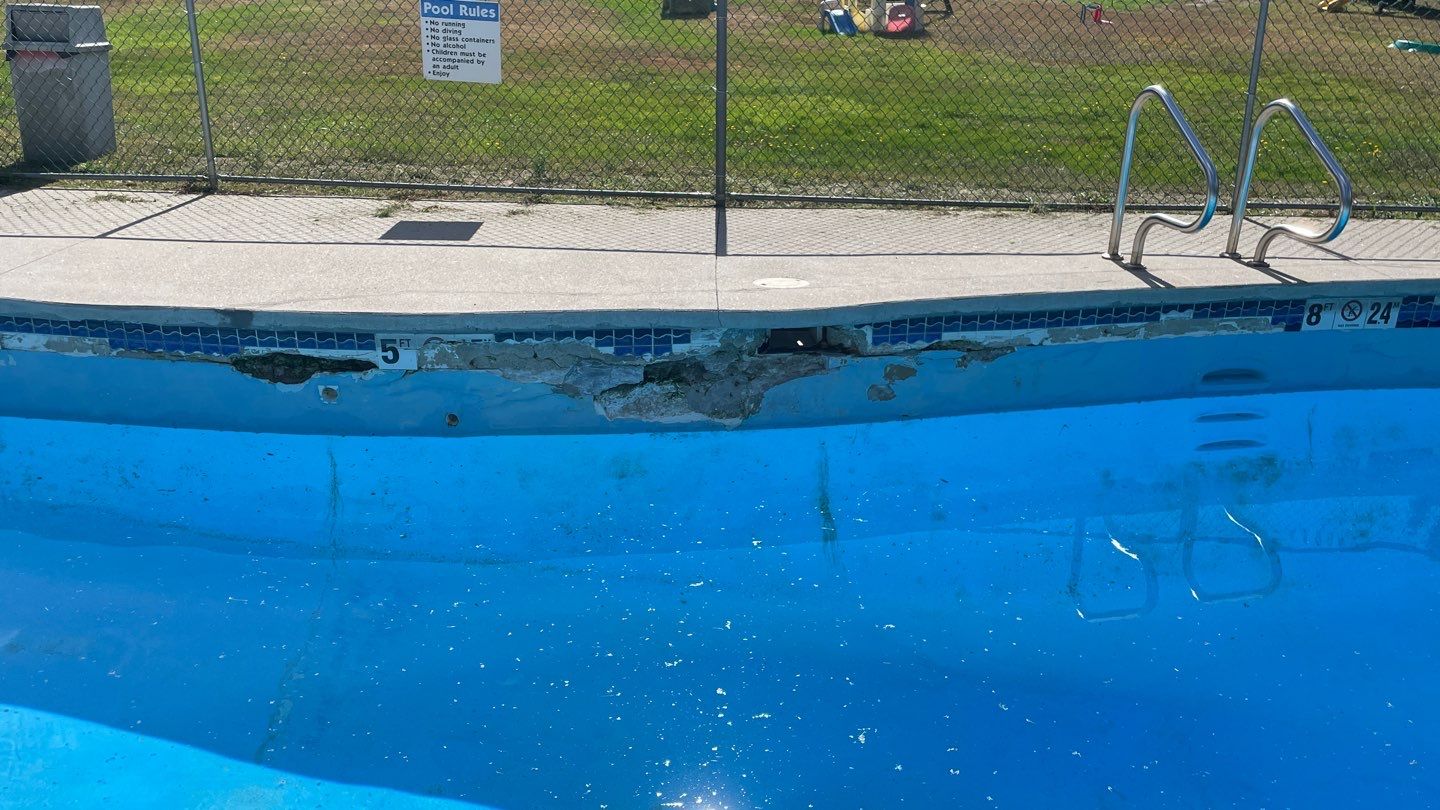 5 A public swimming pool with visible damage and crumbling concrete along the pool edge, near the depth markers, beside a metal pool ladder.