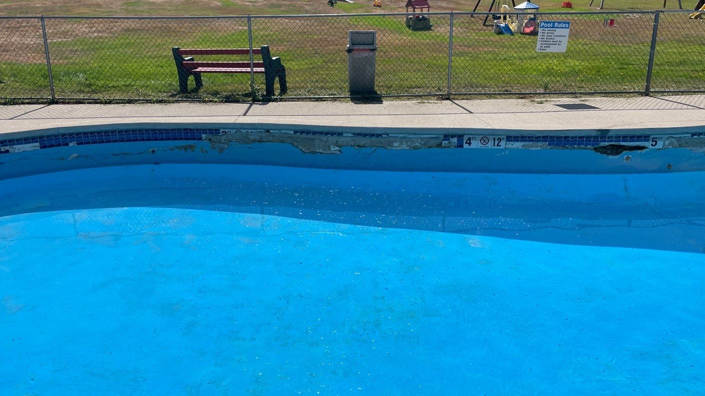 6 Empty outdoor swimming pool with chipped paint and visible damage along the edge; a fence, bench, and sign are visible in the background.