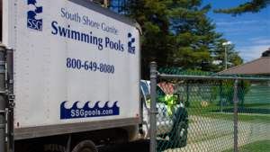 A truck with "South Shore Gunite Swimming Pools" and contact info, highlighting their shotcrete vs gunite expertise, is parked near a chain-link fence by a swimming pool facility and trees.