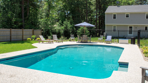 Backyard with an in-ground swimming pool—considering shotcrete vs gunite construction—lounge chairs, a table with an umbrella, green lawn, and a two-story house in the background.