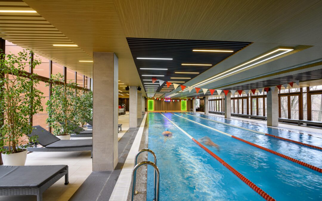 Indoor swimming pool with lane dividers, lounge chairs, potted plants, and large windows; a few people are swimming in the lanes, enjoying the benefits of regular pool maintenance.
