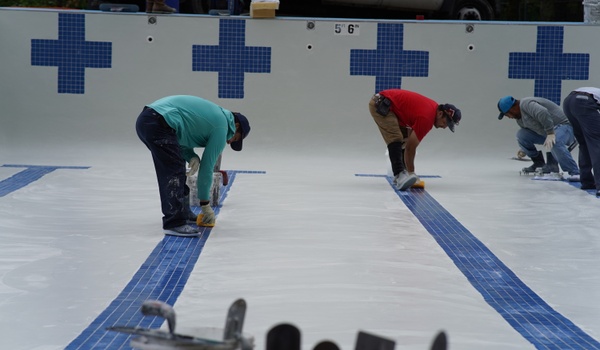 Four workers clean or work on the tiled floor of an empty swimming pool, which has blue striping and cross patterns on the walls.